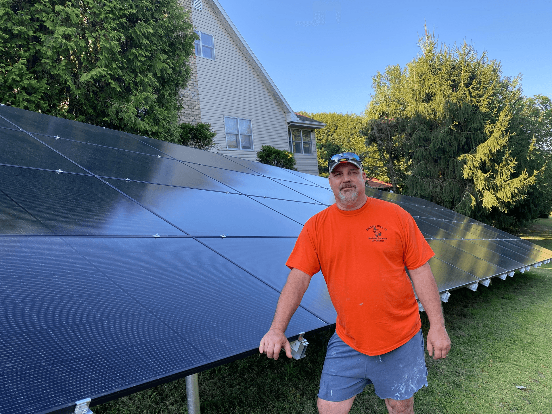 A man in an orange T-shirt and cap stands beside ground-mounted solar panels.
